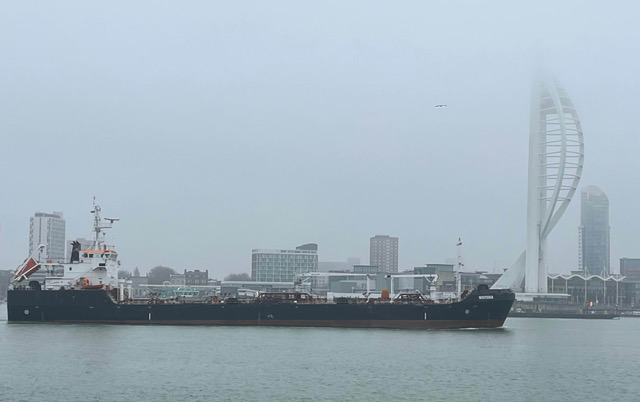 A cargo ship navigating through a foggy harbor with modern city buildings and a distinctive spiral structure in the background.